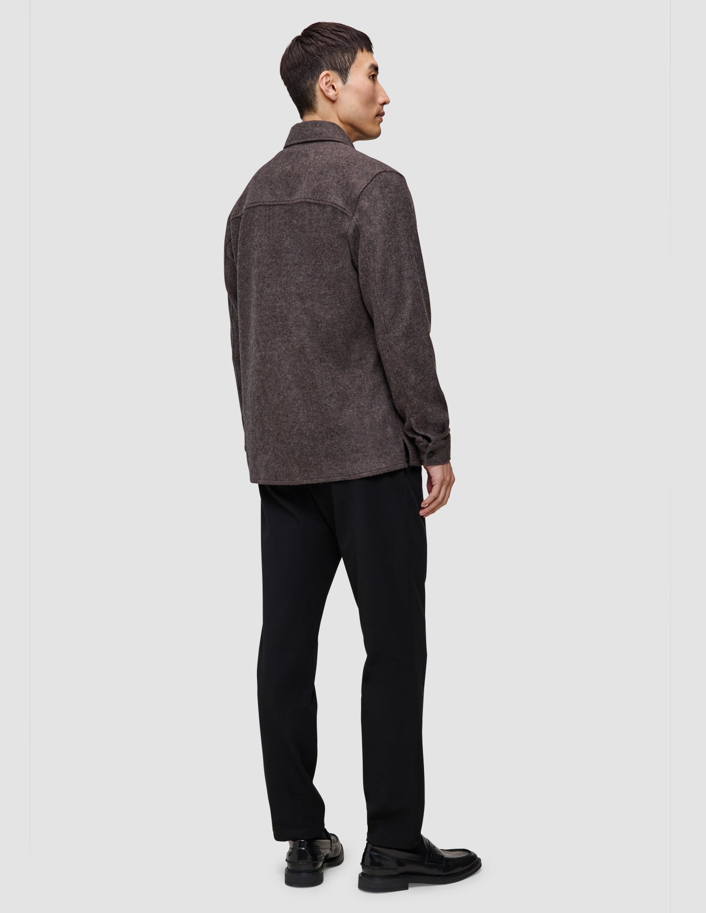 En person set bagfra, iført en dobbeltlags uldskjorte i sadelbrun, sorte bukser og sorte loafers.